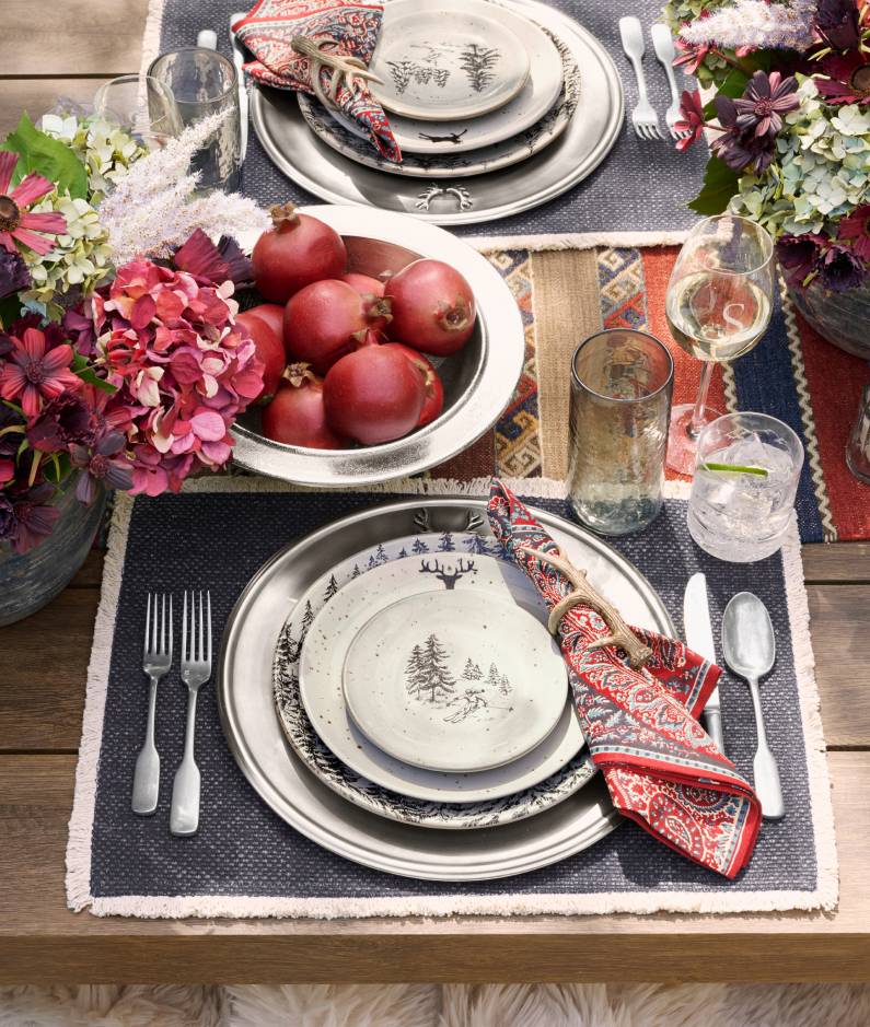 Festive table setting with stacked plates featuring winter forest designs, paired with red paisley napkins. A centerpiece bowl holds red pomegranates, surrounded by vibrant floral arrangements. Silver cutlery and clear glassware accompany the setting on a dark placemat with a colorful woven table runner.