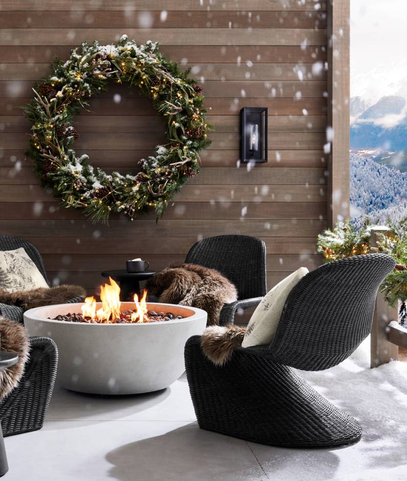 Cozy outdoor seating area with black wicker chairs around a round concrete fire pit. The chairs are draped with faux fur throws, and a large holiday wreath hangs on the wood-paneled wall behind them. Snow gently falls, creating a winter wonderland atmosphere.