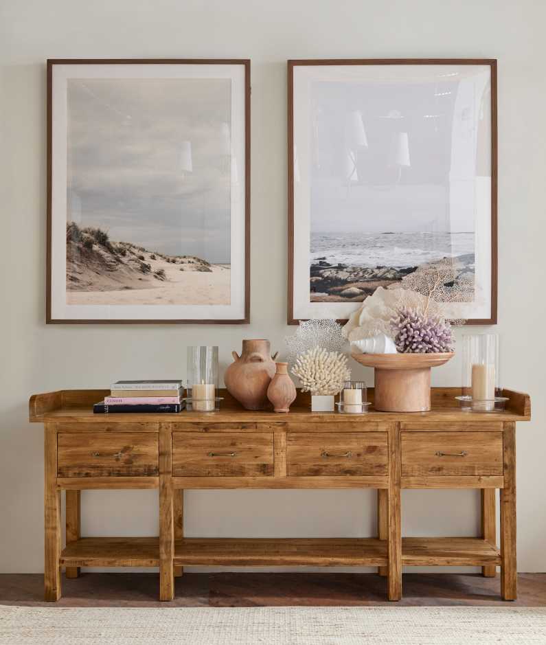 Rustic wooden console table with three drawers and an open shelf, decorated with ceramic vases, candles, and coral pieces. Above the table, two framed beach photographs hang on the wall, depicting serene coastal scenes.