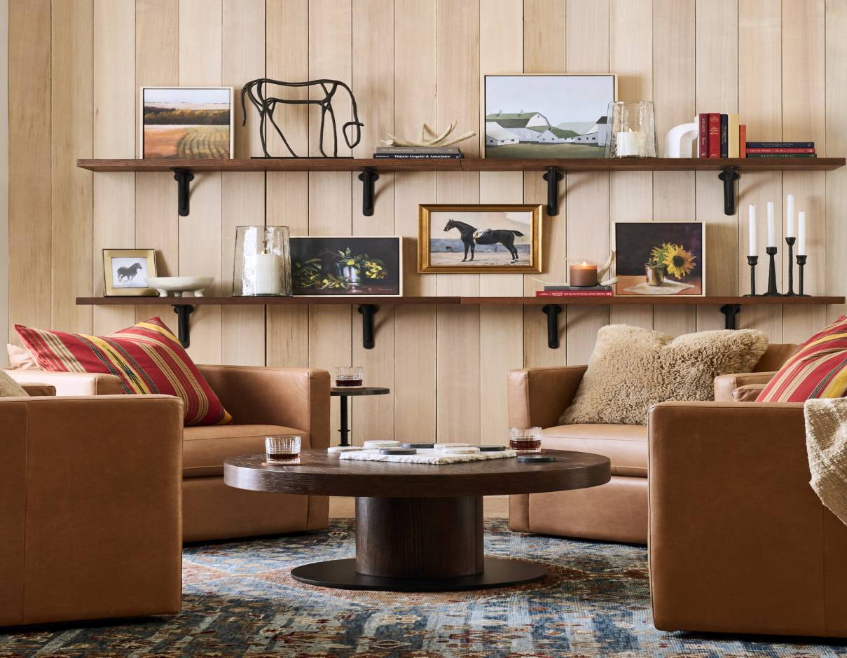 A cozy seating area featuring two tan leather armchairs with striped and fur pillows, centered around a dark wood round coffee table. The light wood-paneled wall behind is decorated with floating shelves displaying framed artwork, books, and decorative accents like candles and a metal sculpture.